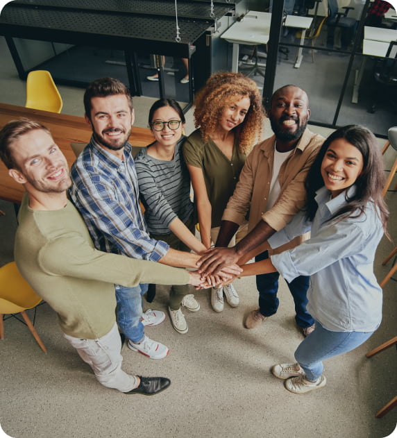 A group of six team members in a creative and collaborative office space with hands in a huddle.